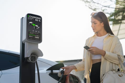 Young woman holding shopping bag and use smartphone to pay for electricity for recharging EV car battery from charging station at city mall parking lot. Modern woman go shopping by eco car. Expedient