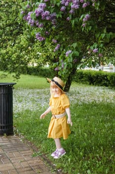 A little girl in a yellow dress and straw hat wearing a bouquet of lilacs. A walk in a spring park, blossoming lilacs