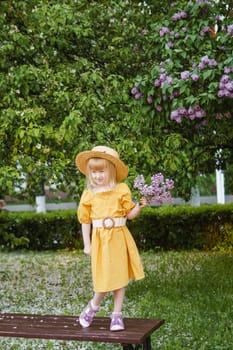 A little girl in a yellow dress and straw hat wearing a bouquet of lilacs. A walk in a spring park, blossoming lilacs