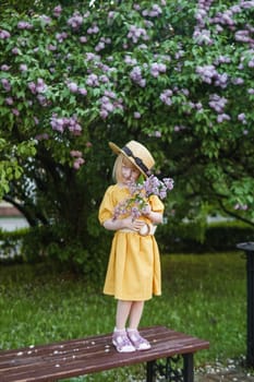 A little girl in a yellow dress and straw hat wearing a bouquet of lilacs. A walk in a spring park, blossoming lilacs