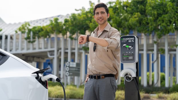 Young man checking time on smartwatch while EV charger to recharging battery from charging station in parking lot. Rechargeable EV car for sustainable eco friendly urban travel. Panorama Expedient