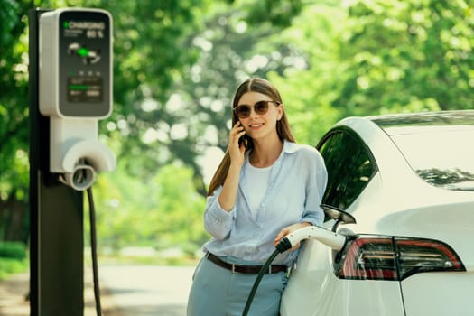 Young woman talking on smartphone while recharging electric car battery charging from EV charging station during vacation holiday road trip at national park or summer forest. Exalt