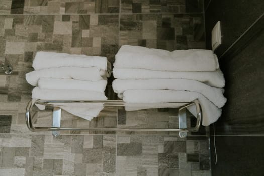 Two stacks of white clean terry towels lie on a metal shelf in a shower with gray tiles, close-up side view.