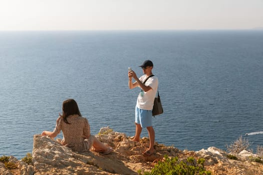 One cool guy in a cap takes a smartphone photo of a girl in a dress sitting on the top of a mountain and looking to the side against the backdrop of a blurry sea on a sunny summer day, close-up side view.