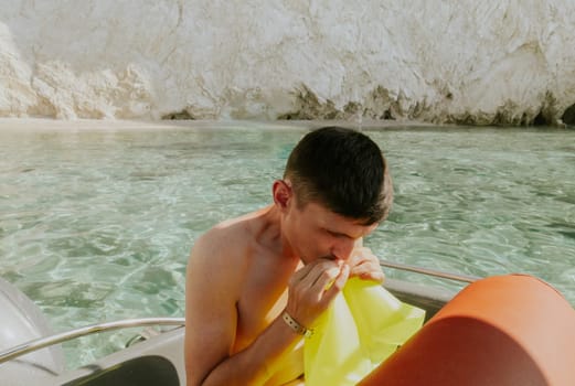 One young handsome Caucasian brunette guy inflates a light green lifebuoy with his mouth while sitting in a boat against the background of blurred rocks on a sunny summer day, side view close-up.