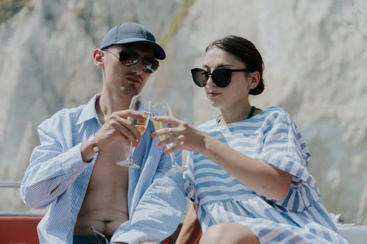 Portrait of one young Caucasian beautiful brunette couple in sunglasses knocking glasses of champagne while sitting in a boat against the backdrop of blurred rocks while sailing on the sea on a sunny summer day, close-up side view.