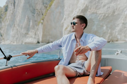 Portrait of one young Caucasian handsome guy in sunglasses sits at the stern of a boat, holds a glass of champagne in his hand, looks away and enjoys sailing on the sea on a private boat on a sunny summer day, close-up side view with selective focus.