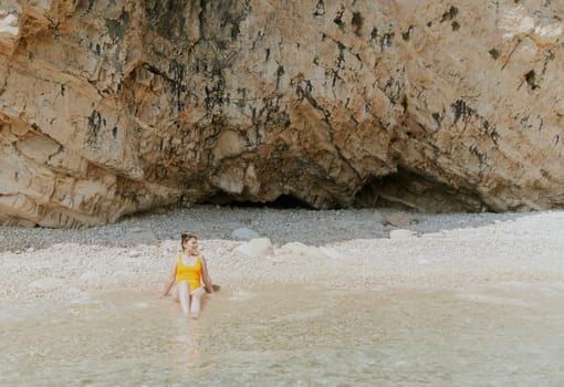 Portrait of one young Caucasian beautiful brunette girl in a yellow swimsuit sits sunbathing and looking to the side on the shore of a rocky deserted beach near the rocks on a sunny summer day, close-up side view.