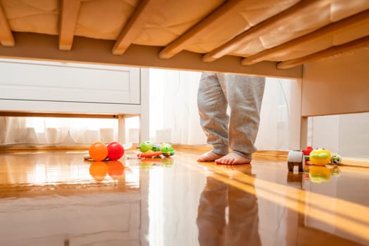 Baby standing near a crib with toys on the floor. View from under the bed. The window brings in