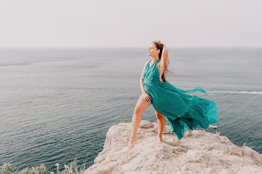 Side view a Young beautiful sensual woman in a mint long dress posing on a volcanic rock high above the sea during sunset. Girl on the nature on overcast sky background. Fashion photo