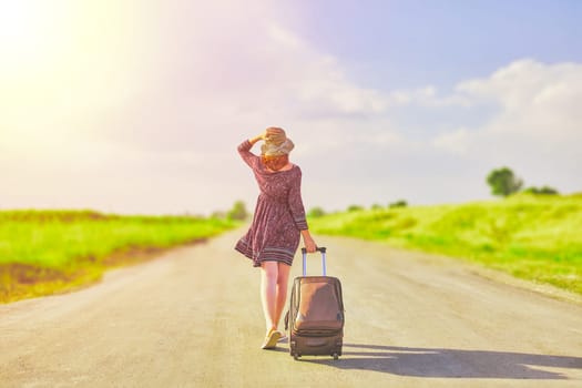 Beautiful woman in summer dress and hat hitchhiking with suitcase at spring. Female walking on the road with baggage at sunny day. Hitchhiker concept