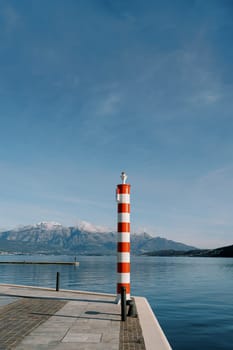 Small striped red and white lighthouse on a pier by the sea against the backdrop of mountains. High quality photo