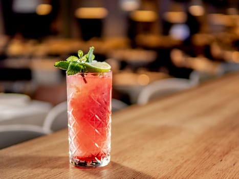 Red non-alcohol cocktail with crushed ice in glass on wooden bar counter in restaurant interior. Gin alcohol cocktail decorated lime and mint. Pink cold alcoholic cocktail in bar or nightclub background. Copy space