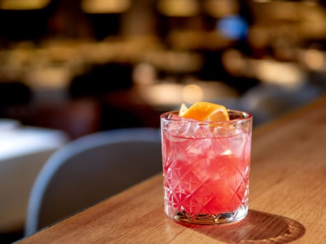 Transparent pink alcohol cocktail with ice, aperol and gin on bar counter in restaurant interior. Alcohol cocktail decorated orange zest spiral. Cold alcoholic cocktail in bar or nightclub. Copy space