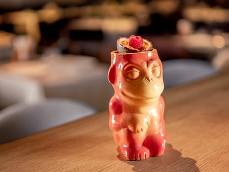 Authentic Tiki Drink Cocktail. Beautiful tropical tiki cocktail on bar counter. Tiki cocktail in monkey mug decorated fig and raspberry on bar in night club or restaurant interior