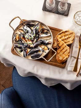 Delicious black mussels in creamy cheese and garlic sauce. Mediterranean black mussels in metal pan on wooden cutting board with grilled bread on restaurant table background, top view