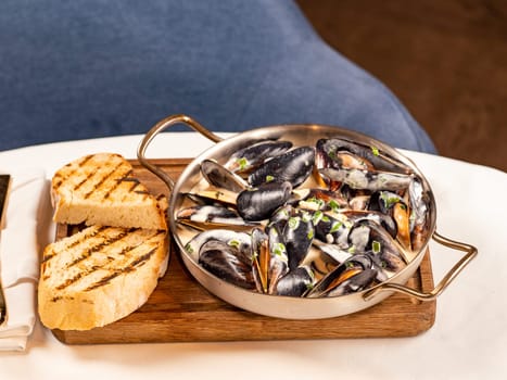 Delicious black mussels in creamy cheese and garlic sauce. Mediterranean black mussels in metal pan on wooden cutting board with grilled bread on restaurant table background