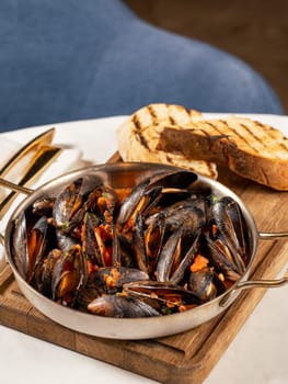 Delicious black mussels in red tomato sauce. Mediterranean black mussels in metal pan on wooden cutting board with grilled bread on restaurant table background
