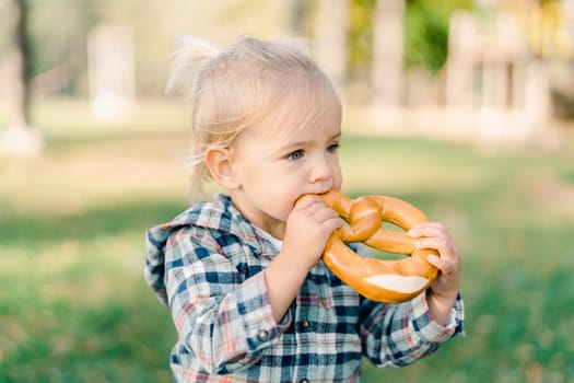 Little girl nibbles a big bagel holding it with both hands on a green lawn. High quality photo