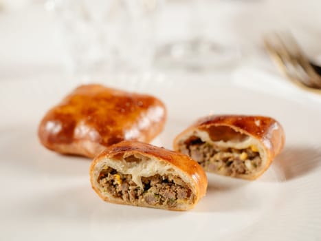 Small russian pies with minced meat Russian Piroshki. Cooked meat hand portioned pies or empanadas cut in half on white plate in elegant restaurant interior. Selective focus, close up.