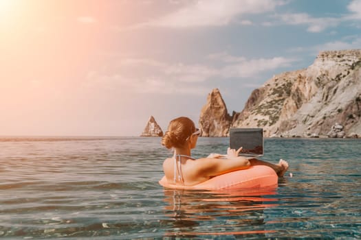 Woman freelancer works on laptop swimming in sea on pink inflatable ring. Pretty lady typing on computer while floating in the sea on inflatable donut at sunset. Freelance, remote work on vacation