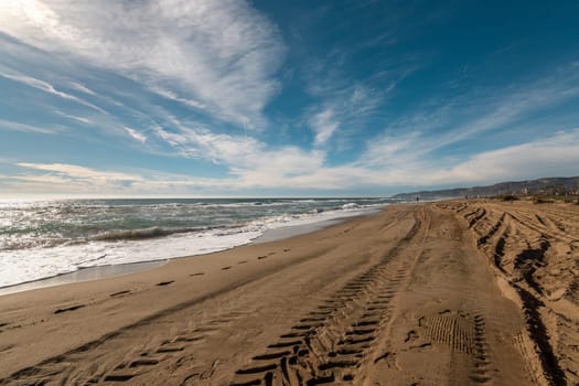 Wide sandy beach with clear tire marks under bright blue sky and wispy clouds, hinting at tranquility.