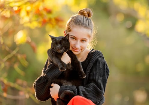 Pretty girl hugging black cat outdoors at street with autumn leaves portrait. Beautiful model teenager with feline animal at park in fall season