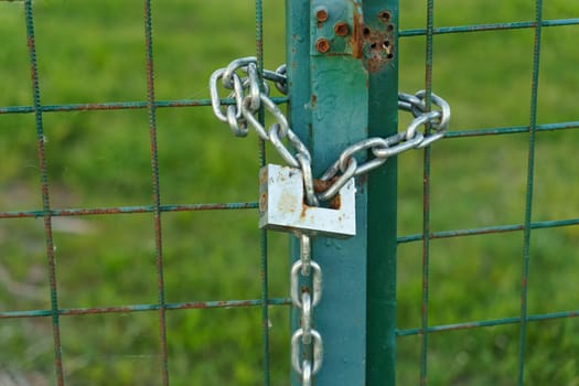 A green gate stands closed with a metal chain securing it shut. The chain is attached to the gates latch, ensuring it remains closed.