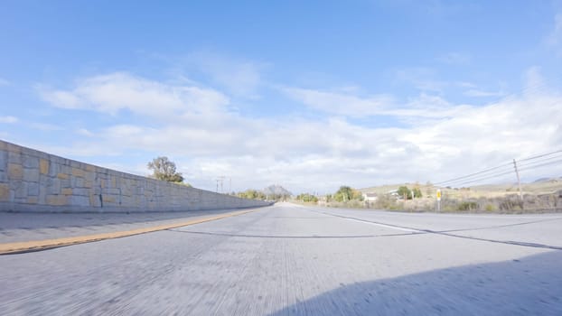 On a crisp winter day, a car cruises along the iconic Highway 101 near San Luis Obispo, California. The surrounding landscape is brownish and subdued, with rolling hills and patches of coastal vegetation flanking the winding road.