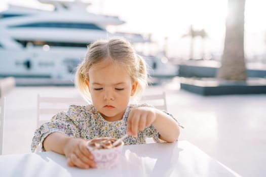 Little girl scoops ice cream from a cup with a spoon, holding it with her hand, sitting at the table. High quality photo