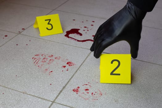 Criminologist hand in black gloves places an evidence marker near bloody footprint on the floor at crime scene.