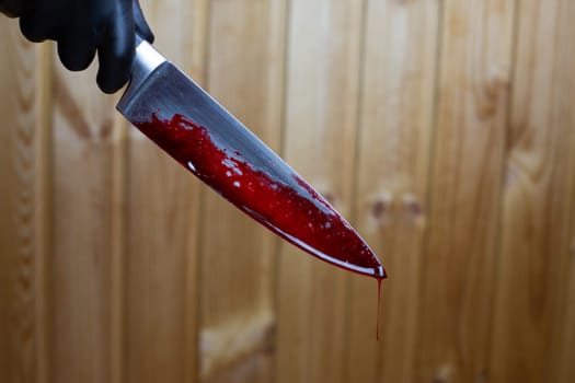 Hand in black glove holds bloody knife with drop of blood flowing down on the background of wooden wall