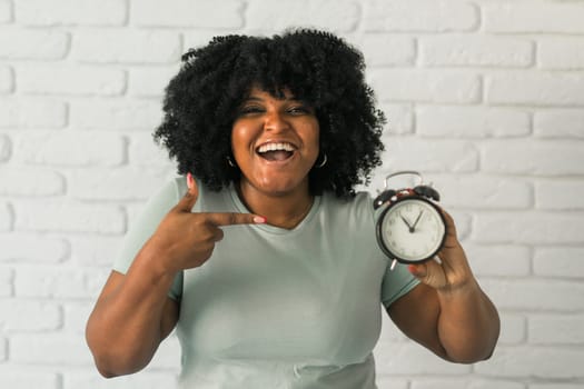 Surprised african american woman holding alarm clock amazed with open mouth and surprise happy face at home, on brick background. Being late and time passing quickly