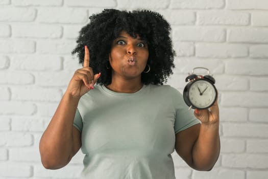 Surprised african american woman holding alarm clock amazed with open mouth and surprise happy face at home, on brick background. Being late and time passing quickly