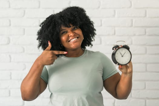 Surprised african american woman holding alarm clock amazed with open mouth and surprise happy face at home, on brick background. Being late and time passing quickly