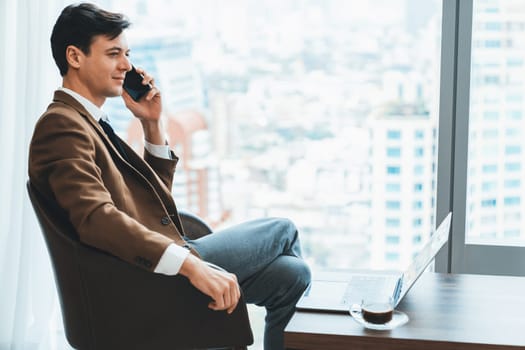 Closeup of handsome businessman making phone call with manager while sitting near window with skyscraper view. Executive manager talking working by using phone and laptop. Look aside. Ornamented.