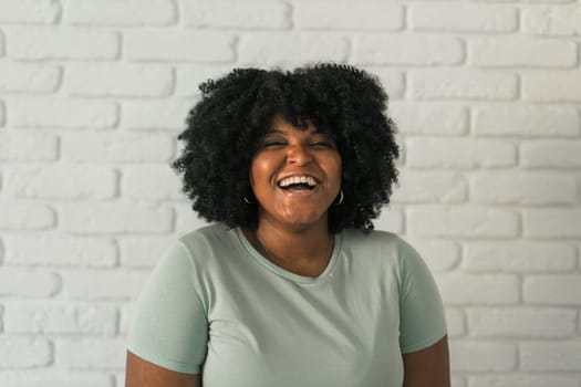Laughing African American woman with an afro hairstyle and good sense of humor smiling and laugh on brick wall at home background. Happiness and good emotions