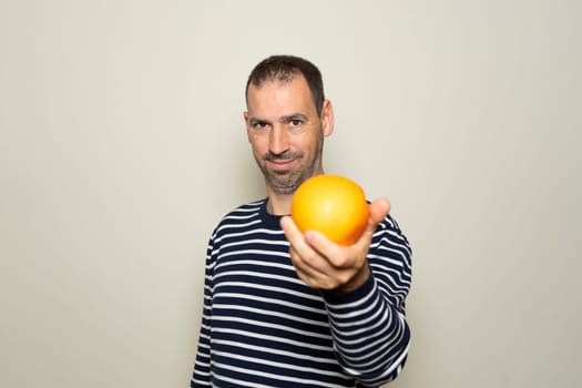 Bearded Hispanic man in his 40s wearing a striped sweater offering you an orange isolated on beige background. Healthy and recommended diet, Mediterranean diet, vitamin C