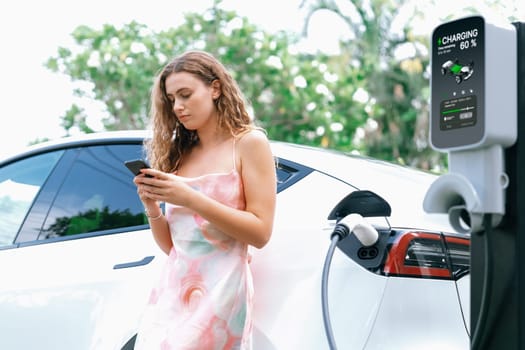 Modern eco-friendly woman recharging electric vehicle from EV charging station. Innovative EV technology utilization for tracking energy usage to optimize battery charging. Synchronos