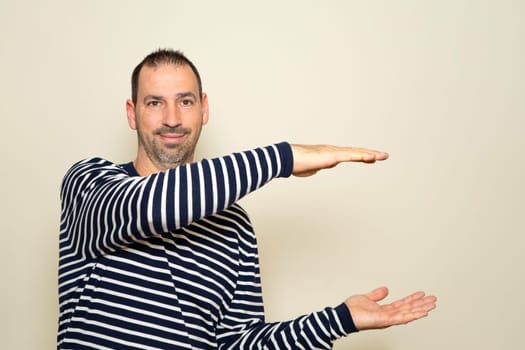Bearded Hispanic man wearing a striped sweater with his arms open in profile offering something while smiling at the camera, isolated on beige background.
