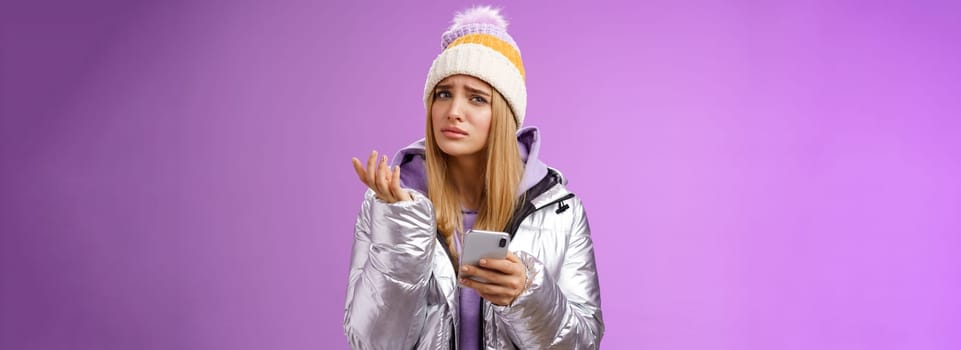 Questioned complicated cute blond girlfriend receive strange message look perplexed confused raising hand shrugging lift eyebrow cannot understand meaning holding smartphone, purple background.