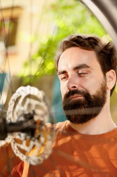 Close-up shot of athletic caucasian man meticulously examining and correcting damages on bicycle wheel chain. Image showing young male cyclist checking tire as yearly bike rubber maintenance routine.