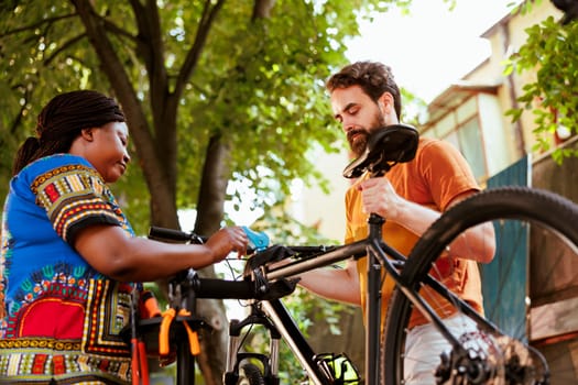 Healthy african american lady assisting her caucasian boyfriend fix his bicycle outside. Active young black woman fastening bike to repair-stand for athletic man to repair using expert tools.