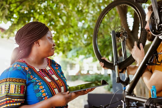 Active black woman enjoys browsing internet while active man fixes and maintains bicycle. Smiling african american female holding digital device as sport-loving caucasian male inspects bike tire rubber for damages.