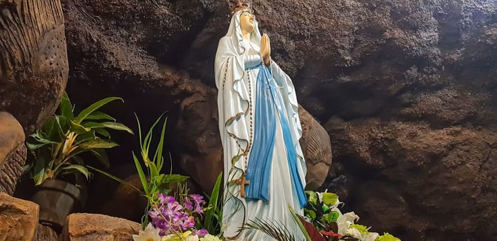 Statue of Holy Virgin Mary in Roman Catholic Church, in the cave of virgin mary, in a rock cave chapel Catholic Church with tropical flowers around in the asian