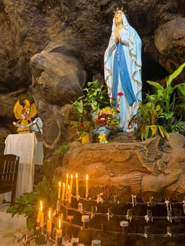 Statue of Holy Virgin Mary in Roman Catholic Church, in the cave of virgin mary, in a rock cave chapel Catholic Church with tropical flowers around in the asian