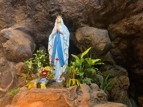 Statue of Holy Virgin Mary in Roman Catholic Church, in the cave of virgin mary, in a rock cave chapel Catholic Church with tropical flowers around in the asian