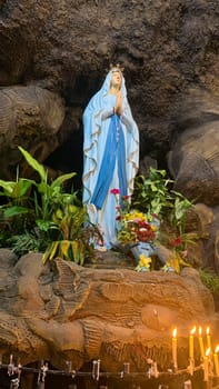 Statue of Holy Virgin Mary in Roman Catholic Church, in the cave of virgin mary, in a rock cave chapel Catholic Church with tropical flowers around in the asian