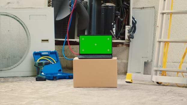 Green screen laptop in front of out of service outside air conditioner. Mock up chroma key gadget display next to damaged external HVAC system in need of expert maintenance