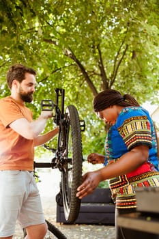 Sporty multiethnic couple inspecting and repairing bike tire and chain in home yard. Healthy and active two persons working outdoors on bicycle maintenance with expert work tool.